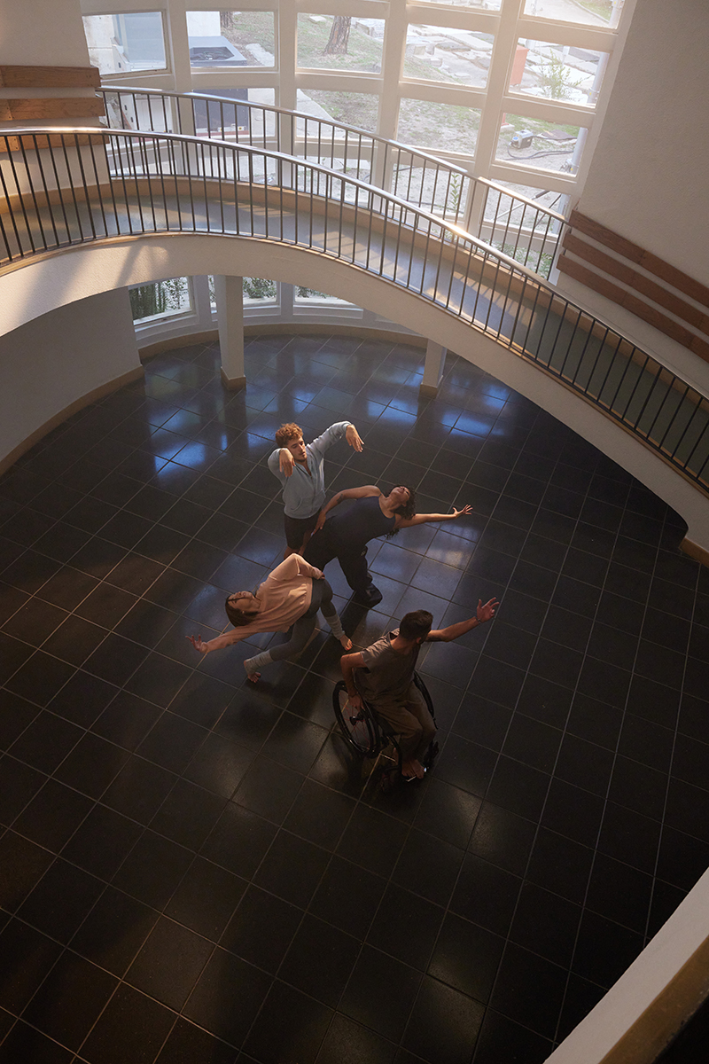 Cuatro bailarines de danza clásica, uno de ellos en silla de ruedas, en un hall rodeado por una escalera caracol