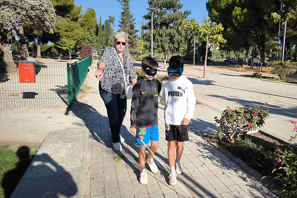 Alumnos en la actividad de Bastón Blanco. Uno de ellos lleva un antifaz y un bastón y el otro unas gafas de simulación. Se ve como una voluntaria de la ONCE les indica por donde ir.