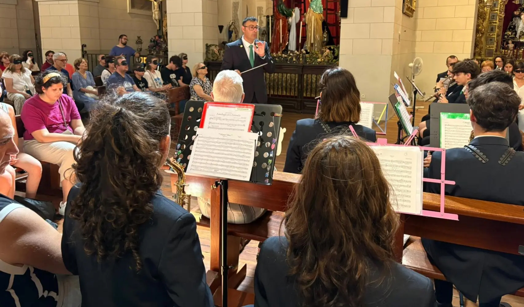 Se ve al director de la banda dirigiendo y a algunos de los asistentes disfrutando del concierto con el antifaz